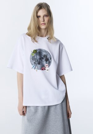 Giovane donna che indossa una t-shirt bianca oversize con una grande luna e piccole toppe colorate, abbinata a una gonna grigia.
