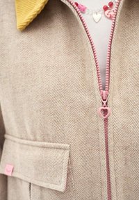 Beežikammandri mantel roosa südamekujulisega tõmblukutrükiga, kollase krae, külgmise taskulapi ja sees nähtava punase südame kaelakeega.