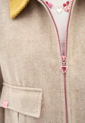 Beżowy płaszcz w jodełkę z różowym suwakiem w kształcie serca, żółtym kołnierzem, klapką kieszeni bocznej oraz widocznym czerwonym naszyjnikiem w kształcie serca wewnątrz.