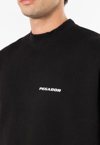 Fekete pamut pulóver, bordás kerek nyakkal és egy kis fehér "PEGADOR" logóval a mellkasán. Simaság jellemzi.