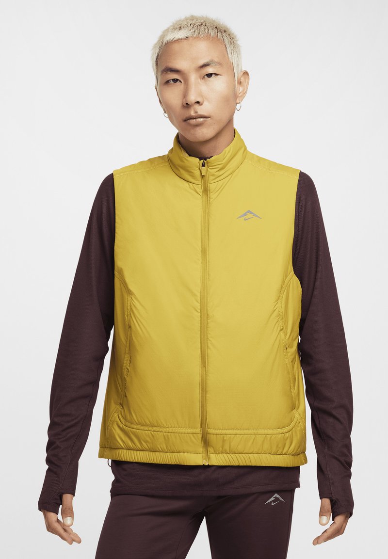 Yellow puffer vest with a high collar, zip closure, and side pockets. Worn over a brown long-sleeve shirt, featuring a logo on the chest.