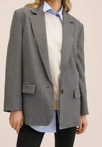 Czarnobiały żakiet w kratę hounds-tooth z wyciętymi klapami, dwoma przednimi kieszeniami, jednoprzyciskowym zapięciem, nałożony na beżowy sweter i niebieską koszulę.