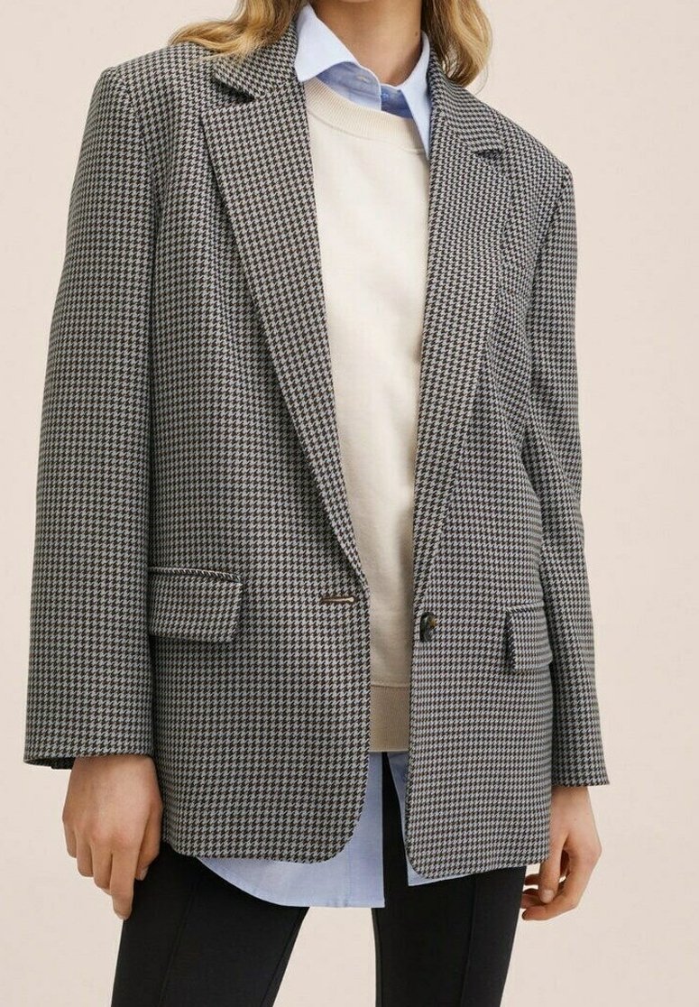 Czarnobiały żakiet w kratę hounds-tooth z wyciętymi klapami, dwoma przednimi kieszeniami, jednoprzyciskowym zapięciem, nałożony na beżowy sweter i niebieską koszulę.