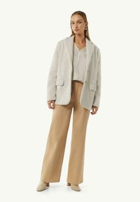 Grijze, gestructureerde oversized blazer met twee voorzakken, gecombineerd met een licht topje en brede beige broek. Neutrale pumps maken de outfit compleet.