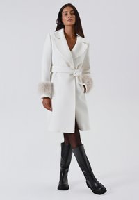 Cappotto bianco con cintura, colletto oversize e maniche in pelliccia sintetica, caratterizzato da una vestibilità sartoriale e una texture liscia. Abbinato a stivali neri fino al ginocchio.
