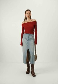 Un pull rouge côtelé à épaules dénudées, associé à une jupe en jean bleu clair avec une fente à l'avant, accessoirisé avec des bottes marron jusqu'aux genoux.