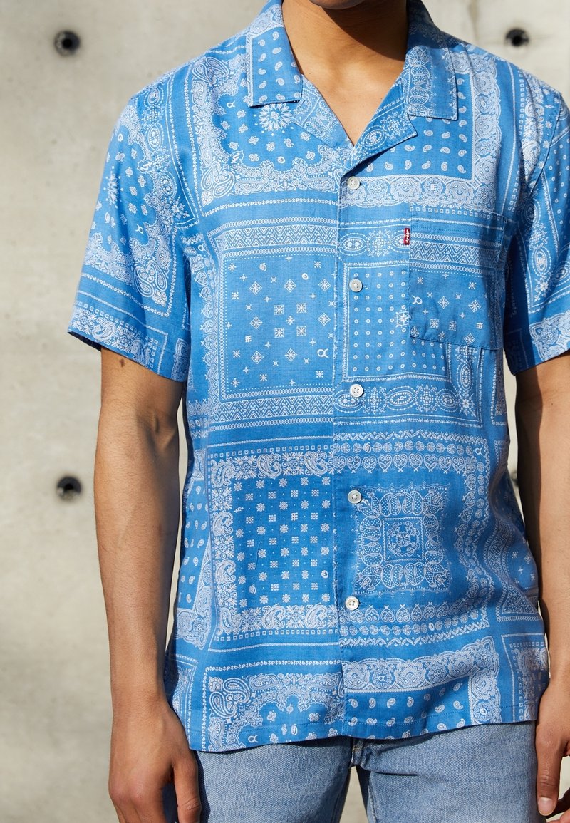 Camicia a maniche corte blu con motivi paisley e geometrici bianchi, il tessuto ha una consistenza liscia, presenta una tasca e bottoni.