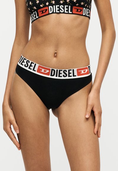 Femme portant des sous-vêtements noirs Diesel avec une ceinture élastique blanche et rouge griffée, debout sur un fond blanc uni.