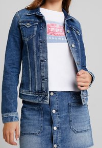 Denim jacket in medium blue; features classic collar, button closure, and chest pockets. Paired with a white graphic t-shirt underneath.