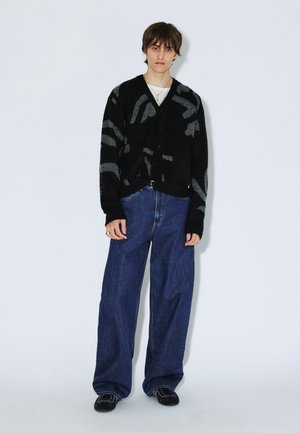 Cárdigan negro con patrón abstracto gris, botones en el frente, usado sobre una camisa blanca. Combinado con jeans de mezclilla azul de pierna ancha y zapatos negros.