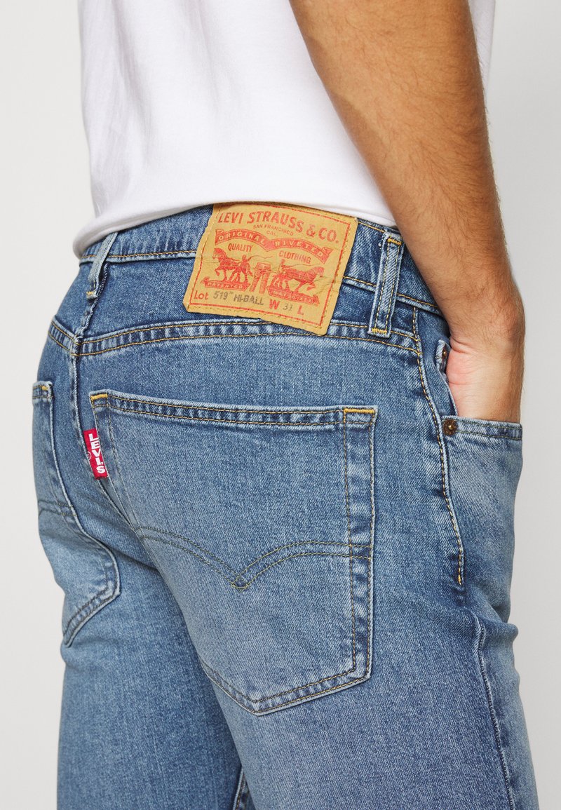 Blaue Denim-Jeans mit geradem Schnitt, ausgestattet mit einem braunen Lederpatch auf der Rückseite und einem roten Etikett. Hell gewaschen mit sichtbaren Nähdetails.