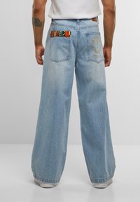 Mann trägt hellblaue weitgeschnittene Jeans mit gesticktem Logo und buntem gemustertem Stoff auf den Gesäßtaschen, steht vor einem einfarbigen Hintergrund.