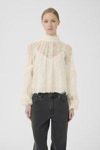 Kvinde iført en gennemsigtig bluse i cremefarvet blonde med høj hals og lange puffede ærmer, parret med højtaljede sorte jeans foran en hvid baggrund.