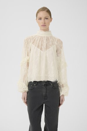 Kvinde iført en gennemsigtig bluse i cremefarvet blonde med høj hals og lange puffede ærmer, parret med højtaljede sorte jeans foran en hvid baggrund.