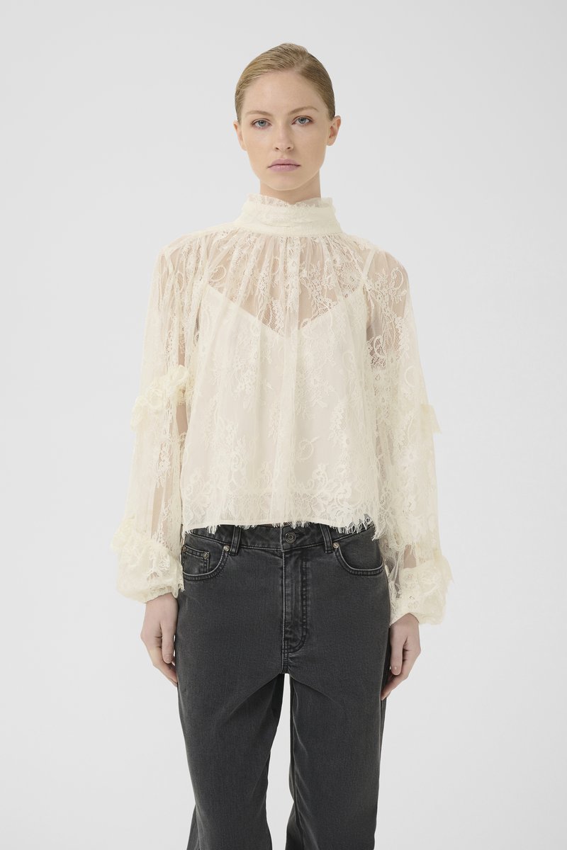 Kvinde iført en gennemsigtig bluse i cremefarvet blonde med høj hals og lange puffede ærmer, parret med højtaljede sorte jeans foran en hvid baggrund.