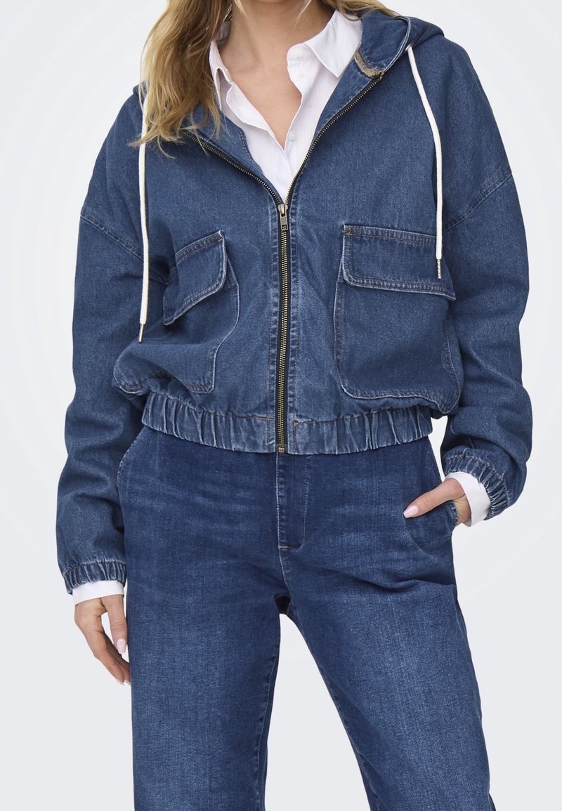 Tmavě modrá džínová bunda na zip s kapucí, dvěma předními kapsami, elastickým páskem a manžetami. Nositá přes bílou košili.