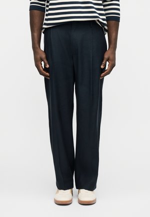 Άνδρας που φοράει μπλε σκούρο παντελόνι με ίσια γραμμή, λευκά αθλητικά παπούτσια και μπλε σκούρη και λευκή ριγέ μακρυμάνικη μπλούζα, στέκεται μπροστά σε μια απλή φόντο.