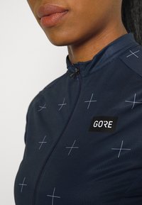 Námořnický cyklistický dres se zipem vpředu. Na hrudi má mřížkový vzor bílých křížů a černé logo „GORE“.