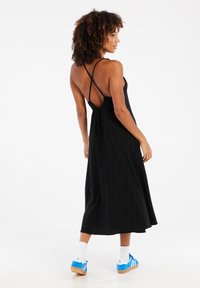 Schwarzes, geripptes, ärmelloses Midikleid mit überkreuzten Trägern im Rücken und einer Bindung. Das Modell trägt blaue Sneaker und weiße Socken.