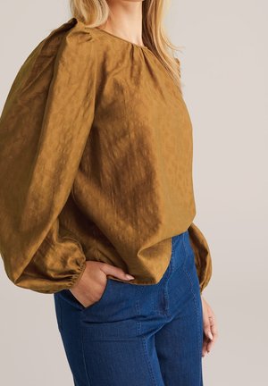 Vrouw draagt een losse bruine blouse met lange puffende mouwen en een high-waisted blauwe spijkerbroek, hand in de zak, neutrale achtergrond.