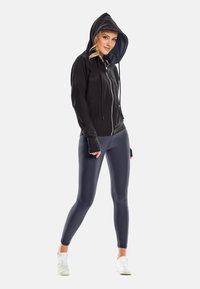 Noor naine musta tõmblukuga kapuutsiga kapuutsiga, hallides retuusides ja valgetes spordijalatsites, seisab jalgadega kõrvuti valgel taustal.