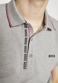 Gray polo shirt featuring a three-button placket, red and black striped collar, and 'BOSS' logo in black on the chest and down the placket.