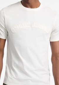 Weißes Baumwoll-T-Shirt mit erhabenem Stickmotiv "GUESS JEANS" in geschwungener Schrift über der Brust. Mit kurzen Ärmeln und rundem Ausschnitt.