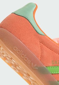 Tenisky Adidas Gazelle s oranžovým semišovým svrškem, zelenými koženými akcenty a průhlednou gumovou podešví s texturovaným gripem.