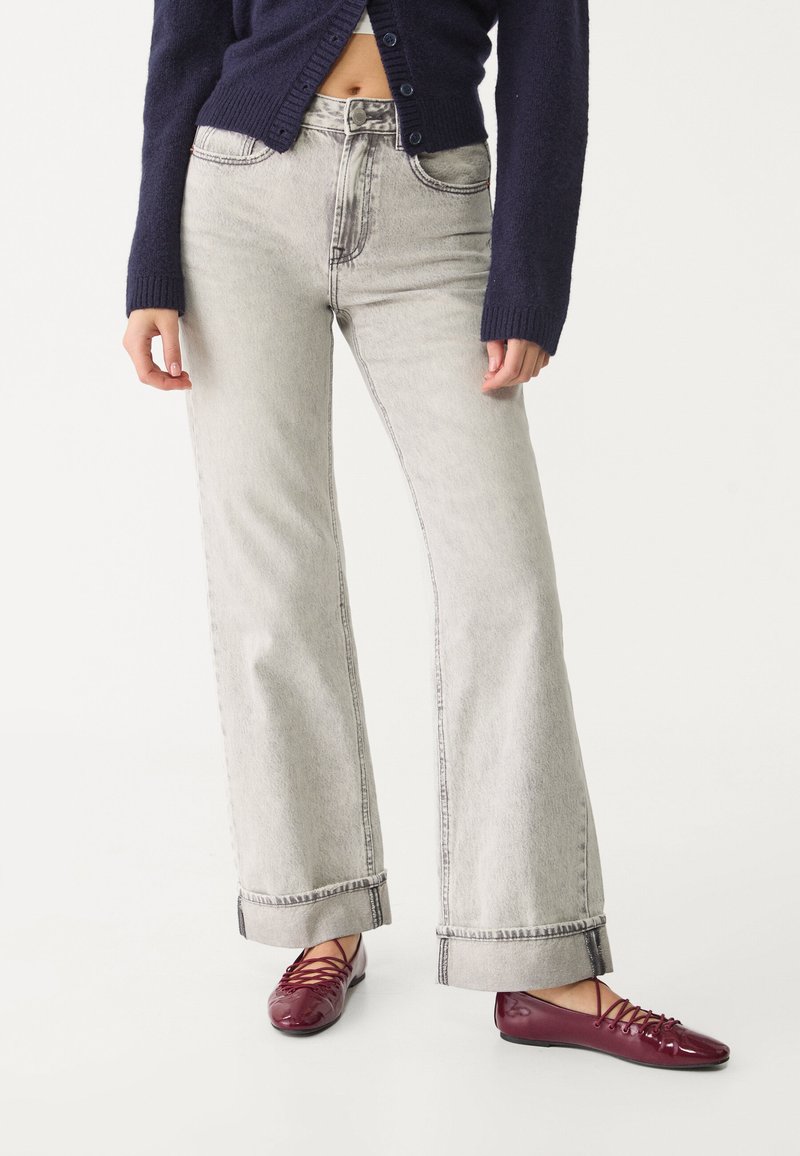 Jeans a gamba larga grigi chiaro con vita alta, caratterizzati da cuciture visibili e orli risvoltati. Abbinati a ballerine borgogna.
