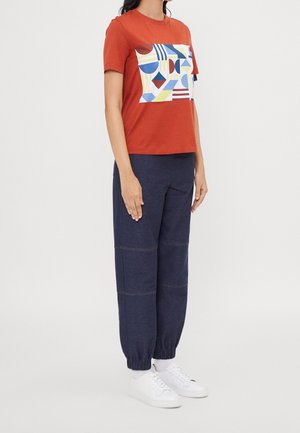 Czerwona bawełniana koszulka z geometrycznym nadrukiem w odcieniach niebieskiego, żółtego i czerwonego; zestawiona z granatowymi spodniami dresowymi z elastycznymi mankietami i widocznymi szwami.