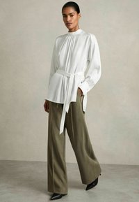 Blusa bianca a maniche lunghe con dettagli plissettati e vita legata, abbinata a pantaloni larghi verde oliva e tacchi neri a punta. Sfondo testurizzato.