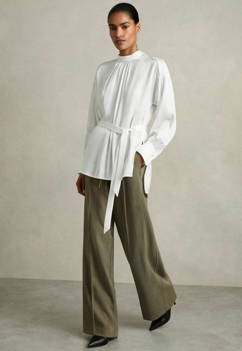 Blusa bianca a maniche lunghe con dettagli plissettati e vita legata, abbinata a pantaloni larghi verde oliva e tacchi neri a punta. Sfondo testurizzato.