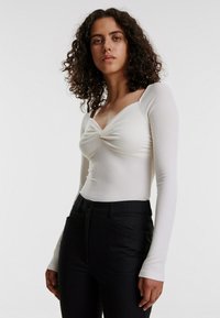 Weißes, geripptes Langarmtop mit gedrehtem Vorderteil, kombiniert mit hoch taillierten schwarzen Hosen, die Taschen und einen maßgeschneiderten Schnitt aufweisen.