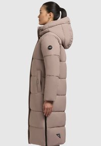 Langer, beigefarbener Puffercoat mit Kapuze, gekennzeichnet durch ein gestepptes Design, einen Reißverschluss vorne und ein Logo-Patch. Seitliche Taschen sind in das Design integriert.