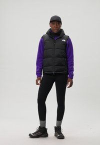 Gilet matelassé noir sur un sweat-shirt violet, associé à des leggings noirs et des chaussettes grises. Portant une casquette noire et des chaussures de randonnée sombres.
