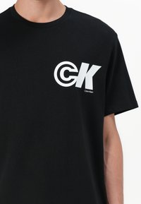 T-shirt de algodão preta com um grande gráfico branco apresentando "CK" e um emblema circular. Gola redonda e mangas curtas, com uma marcação mínima.