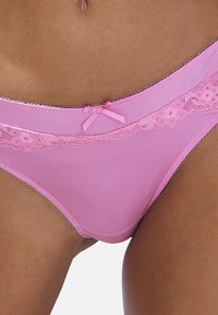 Suurendatud vaade roosa pitsäärisega aluspükstele, millel on lilleline pitsidetail ja väike nool vöökoha peal, kantud päevitusnahal.