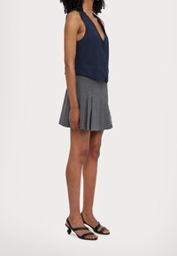 Marinblå ärmlös väst med djup V-ringning och tre knappar, i kombination med en grå plisserad kjol med vertikala ränder och text. Svarta sandaler.