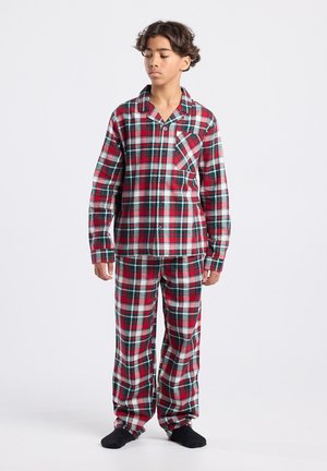 Ein Junge steht da und trägt ein rot-weiß-grünes kariertes Pyjama-Set mit langen Ärmeln und schwarzen Socken vor einem schlichten weißen Hintergrund.
