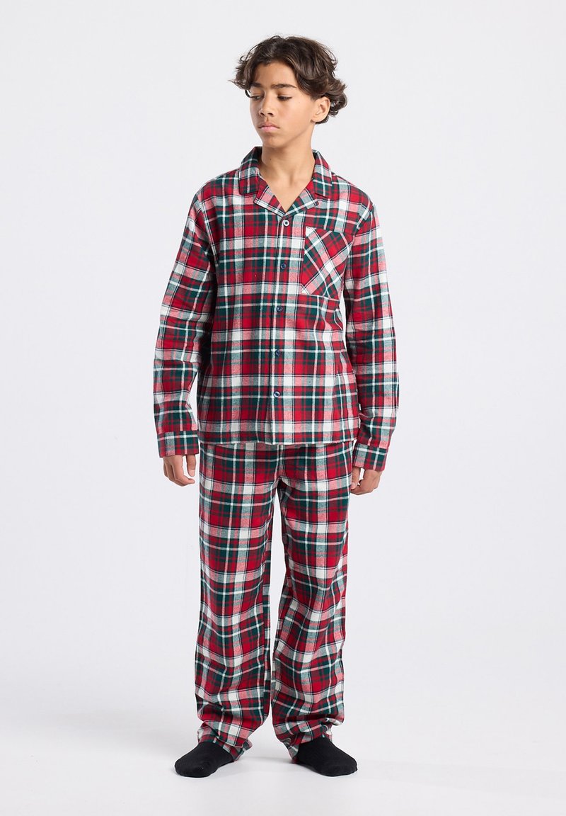 Boy standing wearing red, white, and green plaid long-sleeve pajama set with black socks against a plain white background.