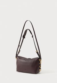 Tumši brūns ādas plecu soma ar regulējamu divu siksnu, zeltainas metāla detaļas fona gaismā.