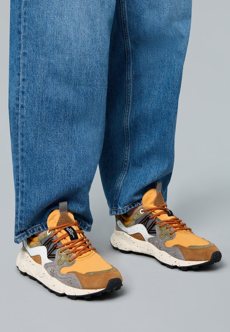 Pestrobarevné tenisky mají kombinaci žluté textilie, šedé a hnědé semišové kůže, skvrnité bílé podrážky a oranžové tkaničky. Nosené s volnými modrými džíny.