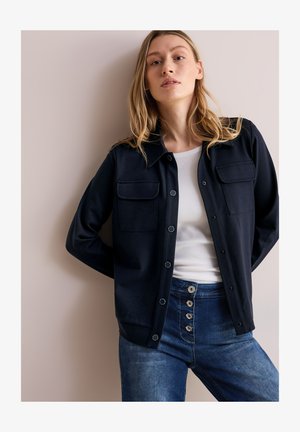 Marineblaues Hemdjackett mit zwei Brusttaschen, aus weichem Stoff. Getragen über einem weißen Oberteil, kombiniert mit hochgeschnittenen blauen Jeans.