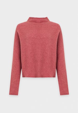 Une femme présente un pull corail, un jean bleu à jambes larges, une ceinture noire et des chaussures plates rouges sur un fond de studio blanc uni.
