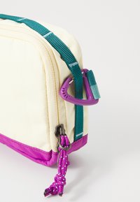 Pochette en toile crème avec une base violette, équipée d'une sangle et de matériels teal. Présente une fermeture éclair et une languette en relief violette.