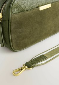Olijfgroene tas met textuur leer en suède panelen, gouden hardware, en een strap met een geweven patroon met twee witte strepen.