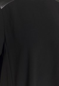 Sort beklædning med en glat, tekstureret overflade og kontrasterende læderlignende skulderdetaljer, der har et simpelt design uden synlige lukninger.