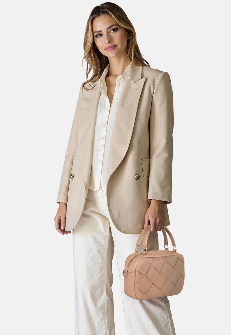 Mujer con blazer beige y pantalones blancos que sostiene un pequeño bolso tejido de color marrón claro sobre un fondo liso y claro.