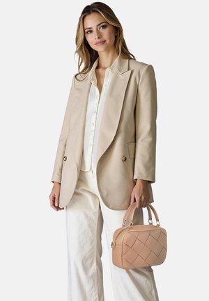 Blazer beige con due bottoni, abbinato a una camicia leggera con bottoni e pantaloni ampi bianchi. Una borsa trapuntata in tono pastello è tenuta in una mano.