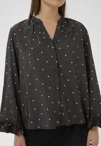 Sort bluse med polka dots, V-udskæring, lange ærmer, rynk ved skuldrene og flæser ved håndleddene. Glat stof med en subtil glans.